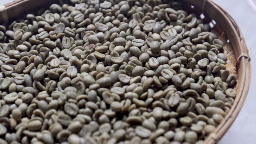 Slow motion video of raw green coffee beans piled inside a traditional woven bamboo basket. Close up shot of unroasted organic coffee seeds resting in a rustic wooden wicker container. Detailed tracking shot of natural raw coffee beans prepared for the roasting process.