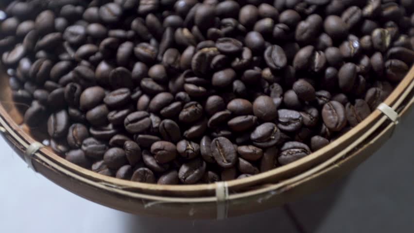 Close up slow motion video of dark roasted coffee beans rotating slowly inside a traditional woven bamboo basket tray. Rich dark brown roasted coffee beans spinning in slow motion within a rustic round wicker sifter under bright studio lighting. Slow motion 50 percent speed shot of a pile of premium roasted coffee grains turning slowly inside a round handwoven wooden container.