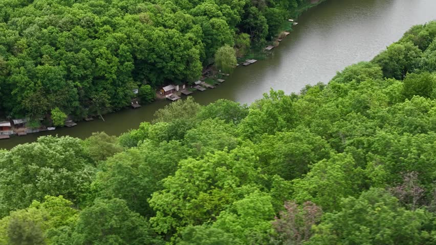 Cinematic aerial drone footage capturing tranquil river winding through dense forest canopy with small rustic wooden cabins and private fishing docks nestled along shore during daytime.