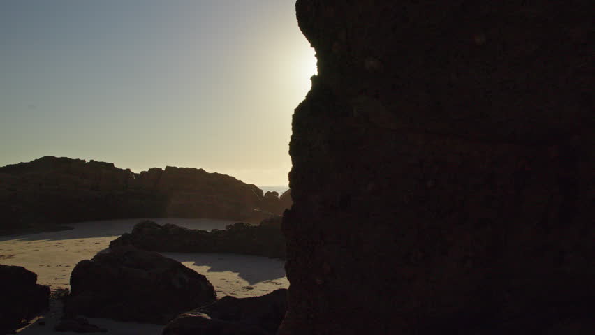 Portugal coastal scene with a bright sunburst beside dark rocks, casting warm light across a sandy beach and sparkling ocean.