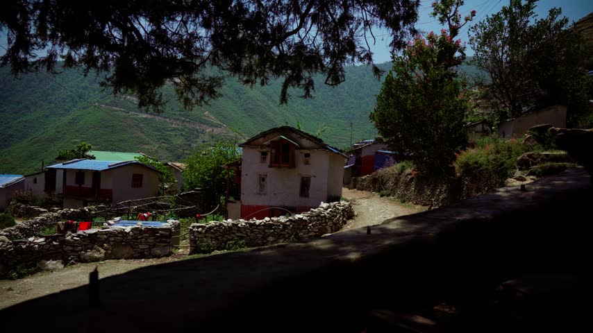 A traditional two-story mud and stone house with a carved wooden window stands beside a stone fence and dirt path in a hill village in Nepal.