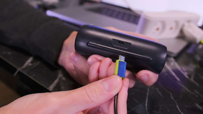 Hand plugging micro USB cable into wireless headphones charging case, close-up of user connecting power cord to portable audio device near laptop workspace. Charging wireless headphones case
