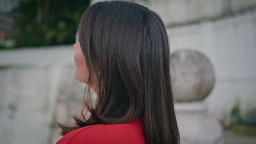 Young woman turning head to camera with gentle smile outdoors. Close up face portrait of confident brunette in red dress near white stone wall, calm happy emotion, feminine grace and natural charm.