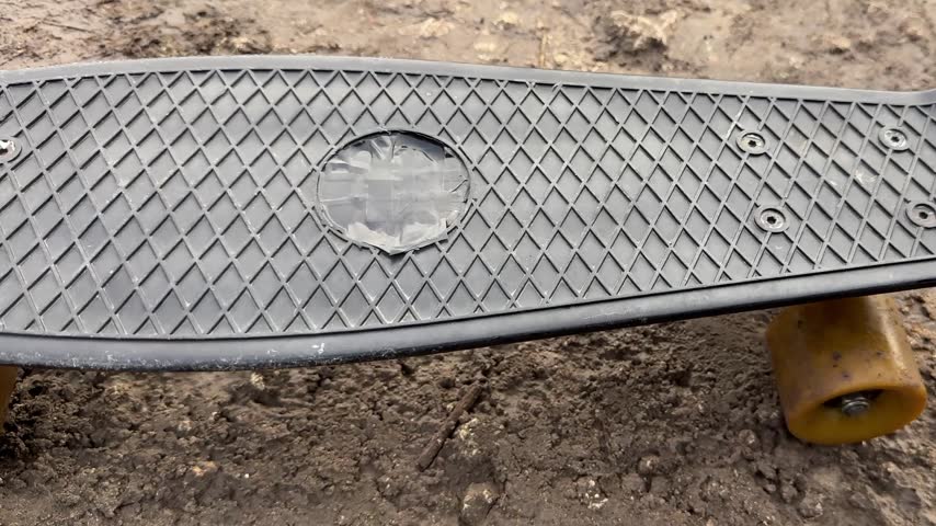 Plastic skateboard deck with single wheel resting on rough dirt surface, low close view showing worn texture, scattered debris, muted outdoor light and empty space