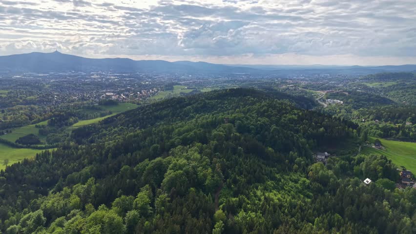 Expansive aerial landscape shows town nestled among rolling hills, dense forests, fields, roads, and farmlands under layered clouds, while sunlight opens across valley and distant mountains