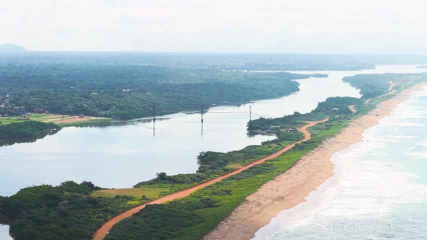 Aerial drone footage of Barra Velha Lagoon and its suspension bridge in Barra Velha, Santa Catarina, Brazil, showing the lagoon, sandy beach, coastal vegetation, pedestrian bridge, and Atlantic Ocean coastline.