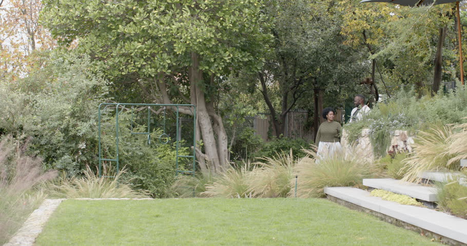 African couple coming from slope, walking down steps and pausing to inspect lawn, copy space. Garden, terrace, duo, pathway, trellis, greenery, shrubs