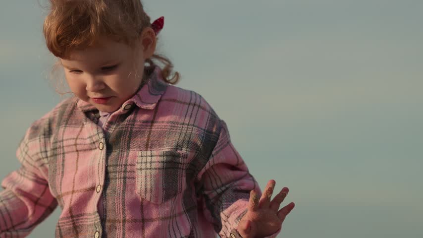 A young child plays outside in a park during sunset. The child wears a plaid shirt and expresses joy while moving around. The background features a clear sky.