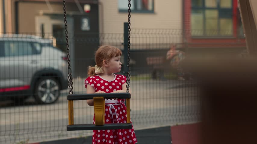 A young girl in a red dress plays on a swing at the park. She watches the surroundings, including parked cars and nearby buildings. It is a bright and sunny day.