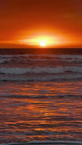 Vertical view of a glowing orange sunset over gentle ocean waves breaking on the shore, with sunlight reflecting on the wet sand.