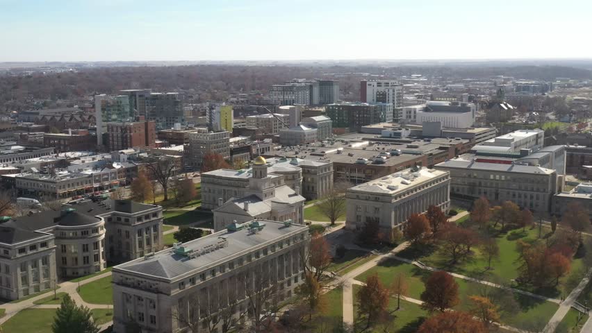 Aerial view of university campus and downtown city skyline during autumn season featuring historic academic buildings golden dome structure and urban cityscape under clear blue sky.