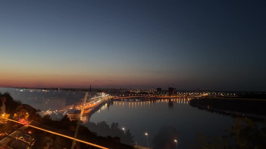 Panoramic evening view of Kyiv and the Dnipro River, Ukraine, with illuminated bridges, city lights, waterfront roads and a sunset sky over the urban landscape.