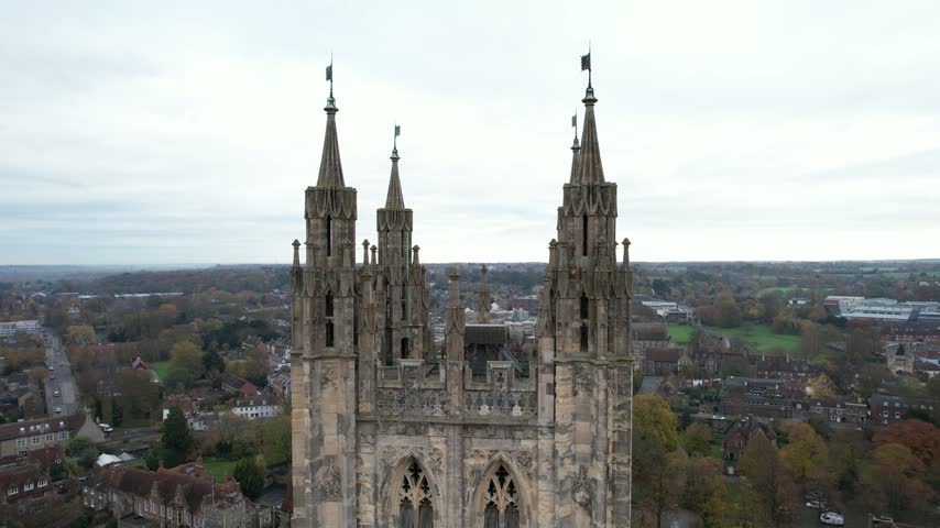 Aerial drone footage captures ornate gothic cathedral spires against a cloudy sky. Cityscape with houses and trees visible below.