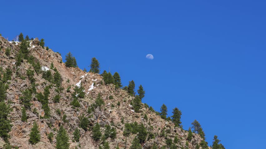 Cinematic view of a rocky mountain slope with evergreen trees and snow patches under a clear blue sky featuring a visible daytime moon.