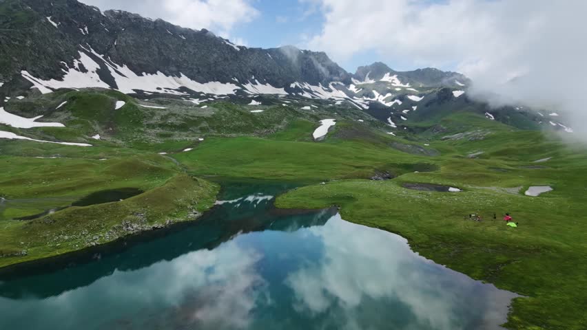 Aerial view of a wild camping site with tents by a turquoise mountain lake under a cloudy sky