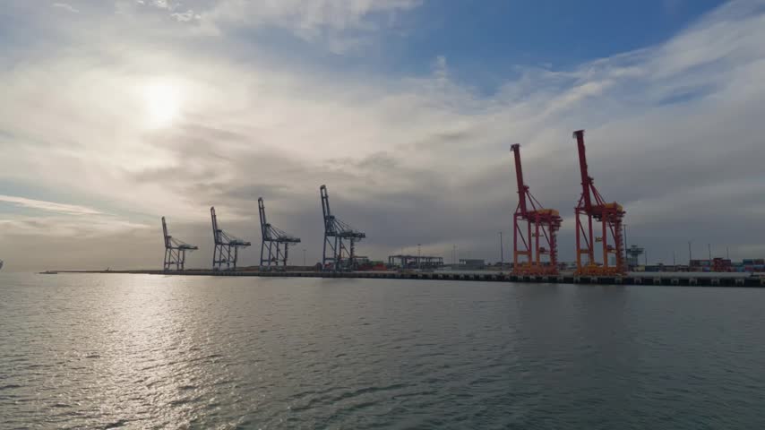 Sunlight glints off calm waters at a bustling port. Large cranes stand tall, ready for cargo. Sky is partly cloudy, with soft light casting long shadows. No people are visible
