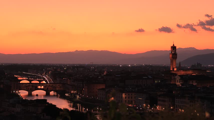 Florence skyline at sunset seen from Piazzale Michelangelo, Italy. Static shot of Ponte Vecchio over the Arno, the Palazzo Vecchio tower and city lights under an orange sky in summer