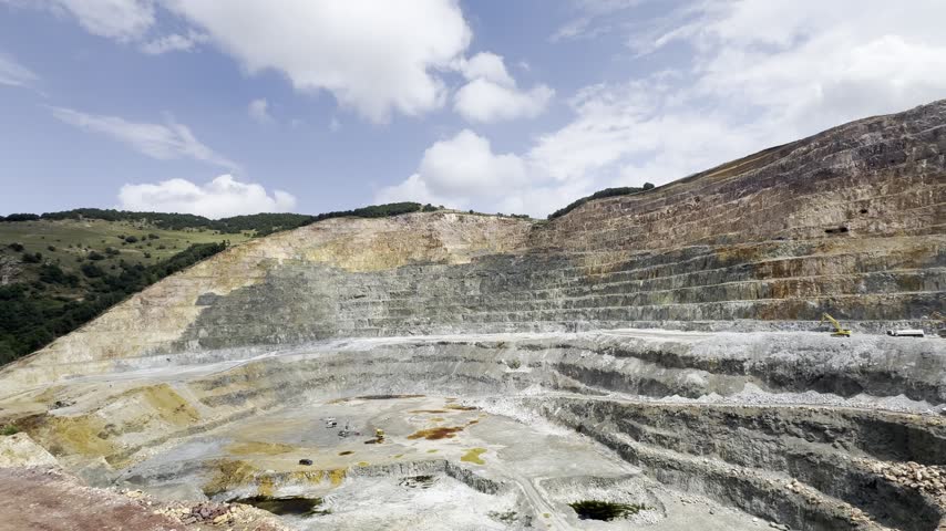 Open-pit quarry with stepped rock walls and exposed geological layers. Heavy excavators are working at the bottom of the quarry, surrounded by rocky terrain, gravel, and puddles. A wide view of an active mining site.
