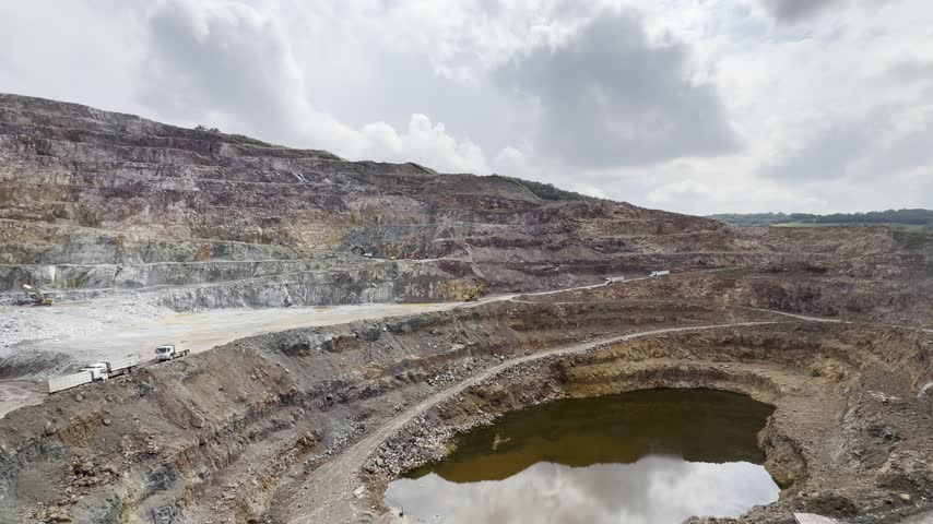 Open-pit quarry with stepped rock walls and exposed geological layers. Heavy excavators are working at the bottom of the quarry, surrounded by rocky terrain, gravel, and puddles. A wide view of an active mining site.
