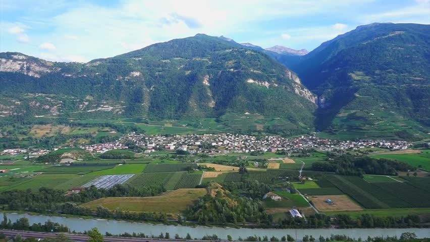 Scenic view of a Swiss Alpine valley with a river, agricultural fields, villages and forested mountains under a partly cloudy summer sky.