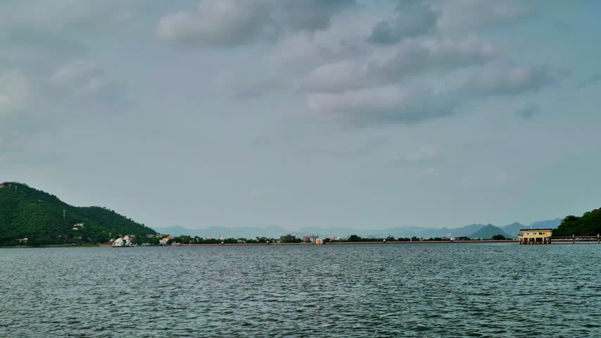 Udaipur�s expansive lake shimmers beneath clouds, framed by green hills, distant Aravalli ranges, a long lakeside promenade, and scattered white architectural structures.