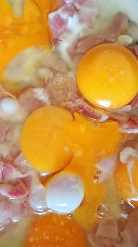 Raw eggs with bright orange yolks cracked into a frying pan on top of fried bacon pieces, egg white starting to set, close-up. Cooking homemade breakfast, fried eggs with bacon, top view.