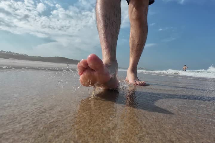 Close up of bare feet walking on wet sand at the beach with ocean waves and blue sky