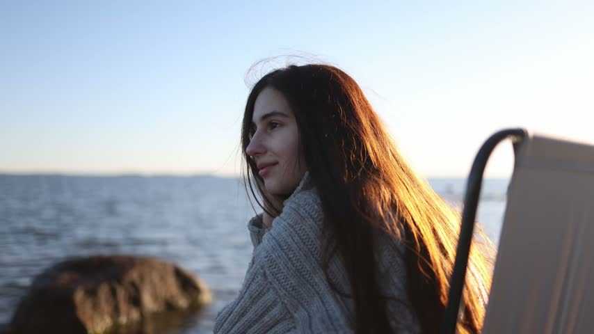 Dark haired woman sitting on a folding camping chair near the sea shown in a close up portrait with a calm coastal atmosphere and natural outdoor setting