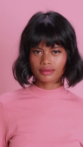 Beautiful Indonesian girl performs mouth zipping motion using fingers to show secret keeping act. Young female with black bob hairstyle smiles after locking lips against pink studio backdrop