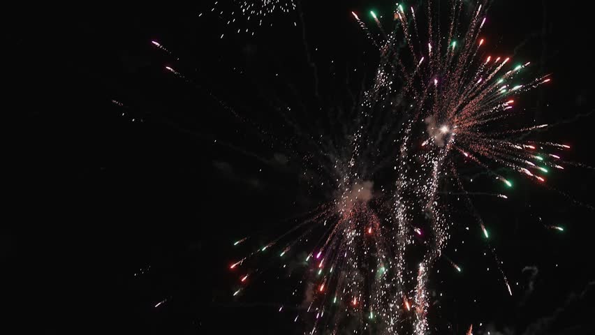 Spectacular fireworks explode across the night sky, sending bright colorful rockets and showers of glowing sparks upward through clouds of smoke. The dynamic display creates a festive atmosphere