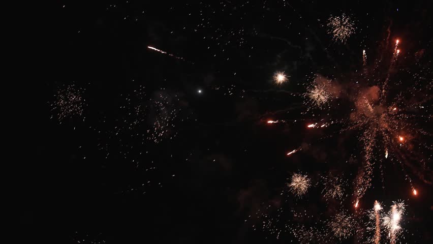Spectacular fireworks explode across the night sky, sending bright colorful rockets and showers of glowing sparks upward through clouds of smoke. The dynamic display creates a festive atmosphere