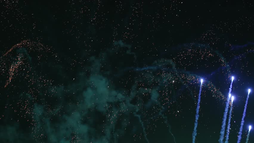 Spectacular fireworks explode across the night sky, sending bright colorful rockets and showers of glowing sparks upward through clouds of smoke. The dynamic display creates a festive atmosphere
