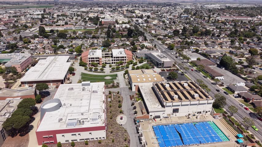 Drone video of a large suburban school campus featuring modern buildings, a bright outdoor swimming pool, and landscaped grounds, set in a residential area under clear daylight. Panning drone shot