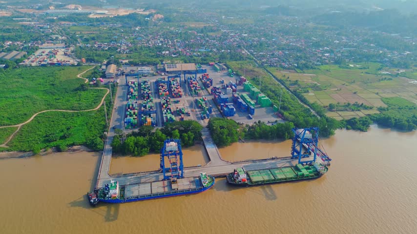 Aerial view of TPK Palaran port terminal with cargo ships, shipping containers, and cranes along the riverbank in Samarinda, East Kalimantan, Indonesia.