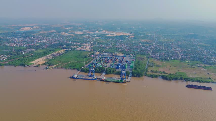 Aerial view of TPK Palaran container terminal on the Mahakam River with cargo ships and cranes, surrounded by lush greenery and urban areas in Samarinda, East Kalimantan, Indonesia.