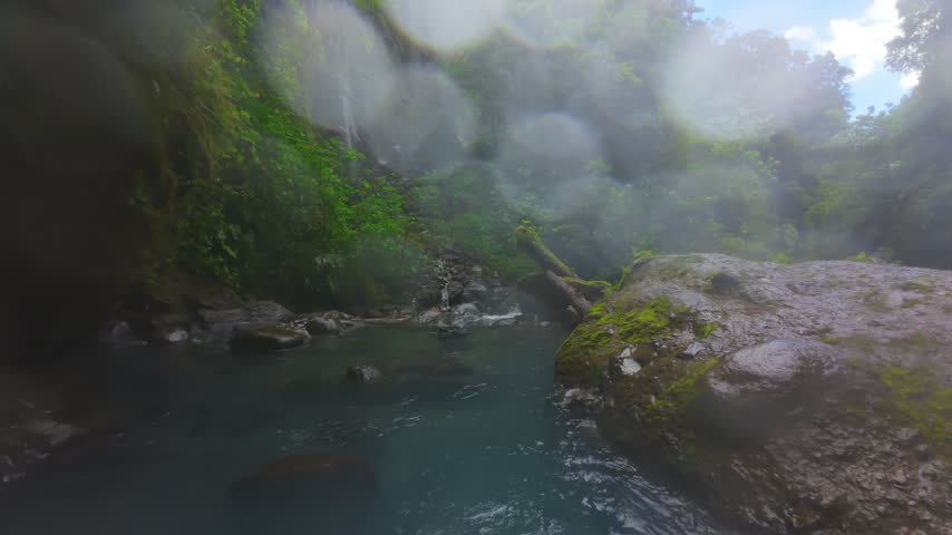 FPV drone flight low over rushing jungle river in Costa Rica with turquoise whitewater rapids mossy boulders tree ferns and mist rising through dense green tropical rainforest canopy.