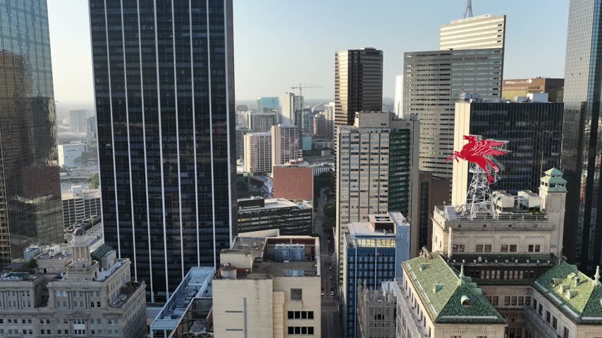 Aerial drone view of dark glass office towers in the Main Street District, Dallas, Texas, at golden hour. Sheer black curtain walls stand over a green tiled roof, low brick blocks and surface parking in the foreground. The small red Pegasus sign turns among them, with tower cranes on the skyline beyond. A lateral tracking pass crosses the blocks.