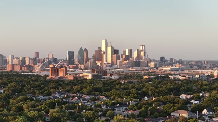 Aerial drone view of the Margaret McDermott Bridge over the Trinity River floodway in Oak Cliff, Dallas, Texas, at golden hour. Two white tied arches with vertical hangers carry Interstate 30 over one wide deck. Mown floodway grass, levees, a hospital campus and dense tree canopy lie behind, with Reunion Tower on the skyline. An orbit reveals both arches.