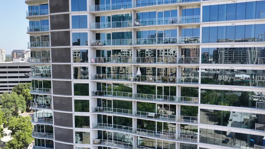 Aerial drone view of a curved glass residence above low roofs in Uptown, Dallas, Texas, at midday. Wavy white balcony edges run the height of the mirrored tower in the foreground. Past its corner are a stone church roof, older houses, parking lots and a rust-framed office block. A tracking shot holds the facade and the blocks behind it.