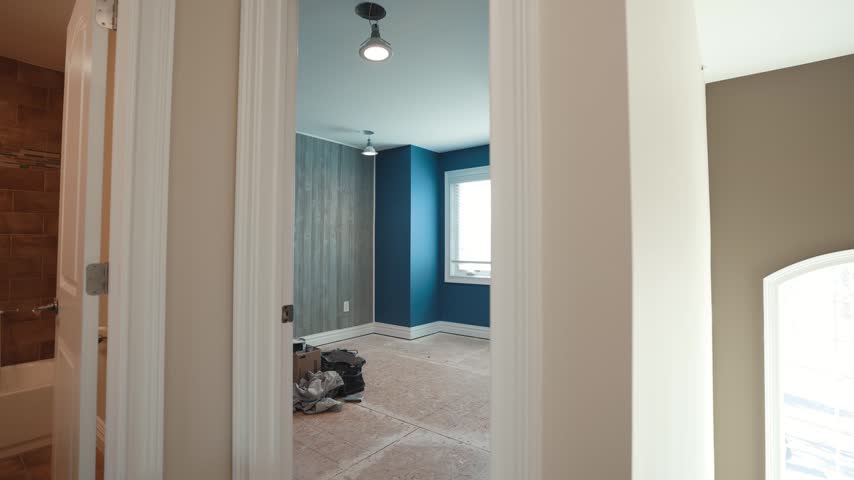 Slow camera motion moving inside small residential bedroom space showcasing deep blue wall paint and decorative wood plank accent wall finishes plus bright window lighting and unfinished flooring.