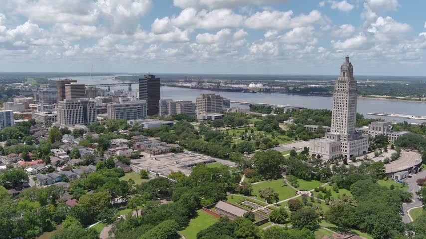Aerial view of Louisiana State Capitol building in Baton Rouge showcasing cityscape green parks residential areas and Mississippi River with industrial background under blue sky.