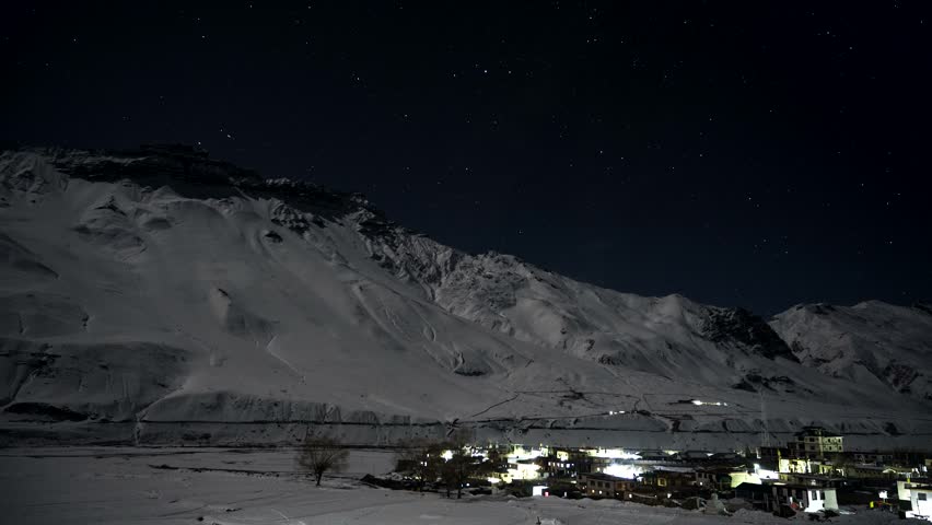 Star trails rotating across night sky above illuminated mountain village in Himalayas. Time lapse video captures celestial movement over vast snow covered peak landscape background.