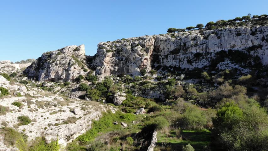 Aerial view moving upwards over rocky limestone cliffs leading to deep open pit quarry in rural Malta. Bright sunny daylight conditions highlight geological formations and local architecture.