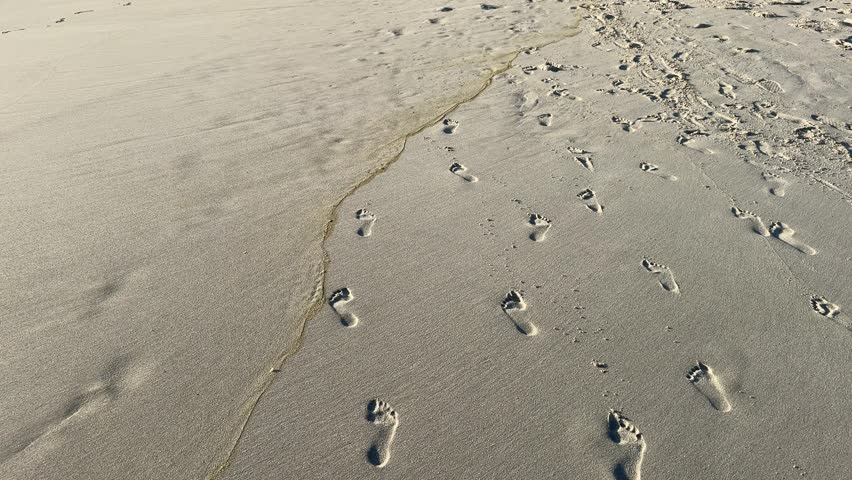 Footprints in sand on ocean beach shore with gentle waves moving over wet sand surface. Cinematic footage showing walking track marks along coastline during calm sunny day.