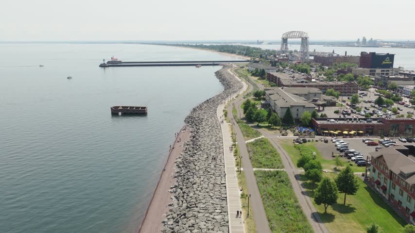 Drone flies over rocky lakewalk breakwater near Canal Park showing Aerial Lift Bridge pier lighthouse and Lake Superior waterfront in Duluth