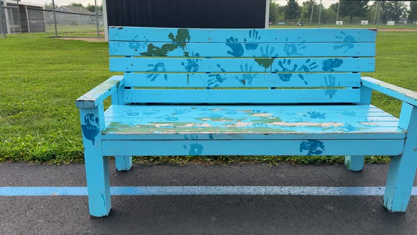 A weathered light blue wooden bench decorated with painted handprints sits beside a sports field, capturing community art and childhood memories outdoors.
