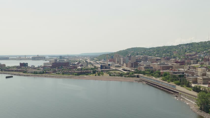Drone flies toward Duluth Lakewalk Beach near Sister Cities Park with smokestack downtown buildings and Lake Superior port on hazy day