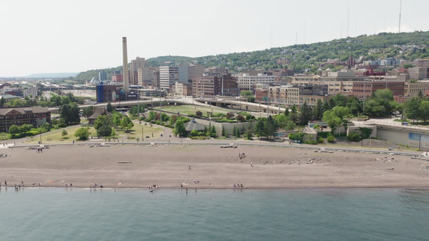 Sandy Lake Superior beach with people walking and lakeside pavilion framed by downtown Duluth skyline and smokestack on hillside