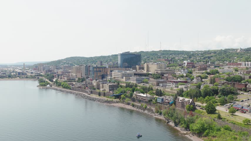 Drone orbits downtown Duluth skyline showing modern glass tower medical buildings and hillside neighborhoods above Lake Superior shoreline