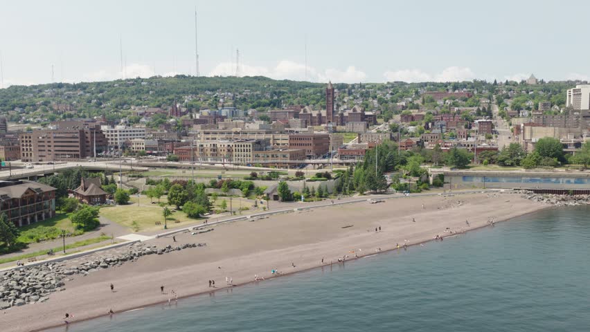 Aerial rising shot of sandy Lake Superior beach with people walking along shoreline and downtown Duluth in background